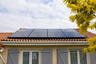 Une maison beige avec un toit recouvert de tuiles canal oranges sur lequel des panneaux solaires sont posés. Arbre avec fleurs jaunes à droite de l'image