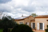 Maison individuelle beige équipée de panneaux solaires et entourée de verdure. Ciel nuageux